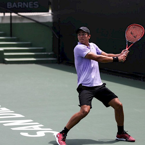 A tennis player in motion, preparing to hit a backhand shot on an outdoor court. The player is wearing a hat, purple shirt, and black shorts.