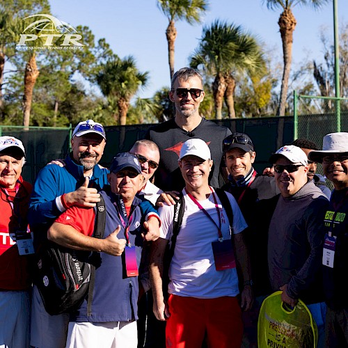 A group of people in athletic wear, some wearing tennis outfits, posing outdoors with palm trees in the background, under a sunny sky, are shown.