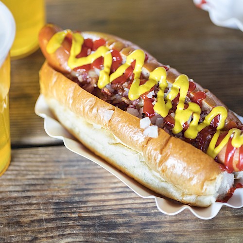 A hot dog with mustard and ketchup and a glass of beer on a wooden table.
