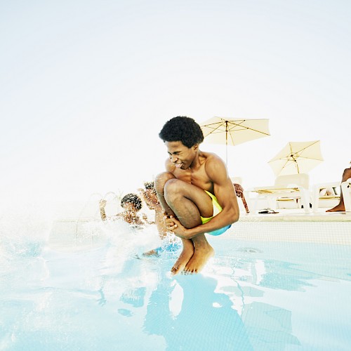 Person jumping into a pool, creating a splash, with others relaxing poolside.