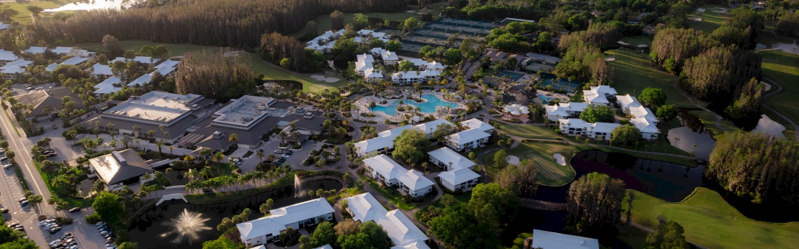 An aerial view of a resort complex with buildings, pools, tennis courts, and surrounding greenery under a partly cloudy sky.