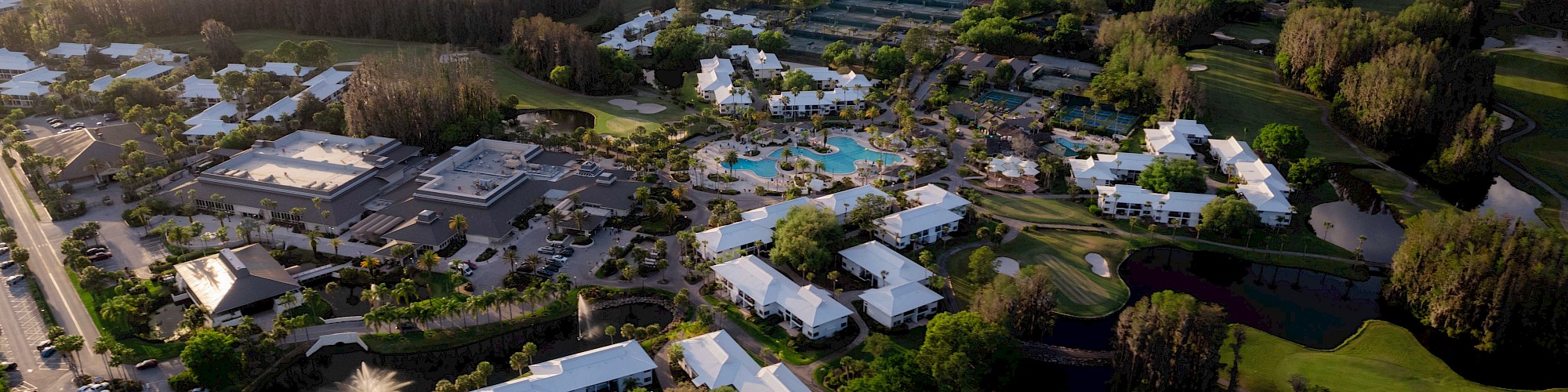An aerial view of a resort complex with buildings, pools, tennis courts, and surrounding greenery under a partly cloudy sky.