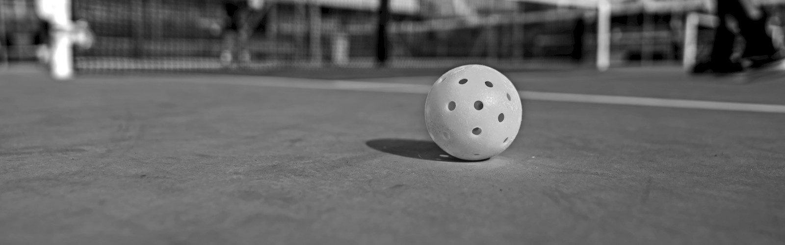 A pickleball sits on a court, with a net and players in the background, suggesting a game in progress.