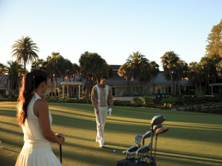 Two people on a golf course at sunset, with palm trees and a large building in the background.