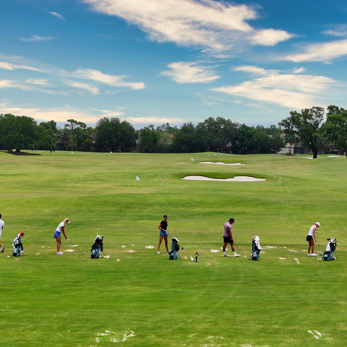 People practicing golf at a driving range, each with their own set of clubs, surrounded by green grass and trees under a partly cloudy sky.