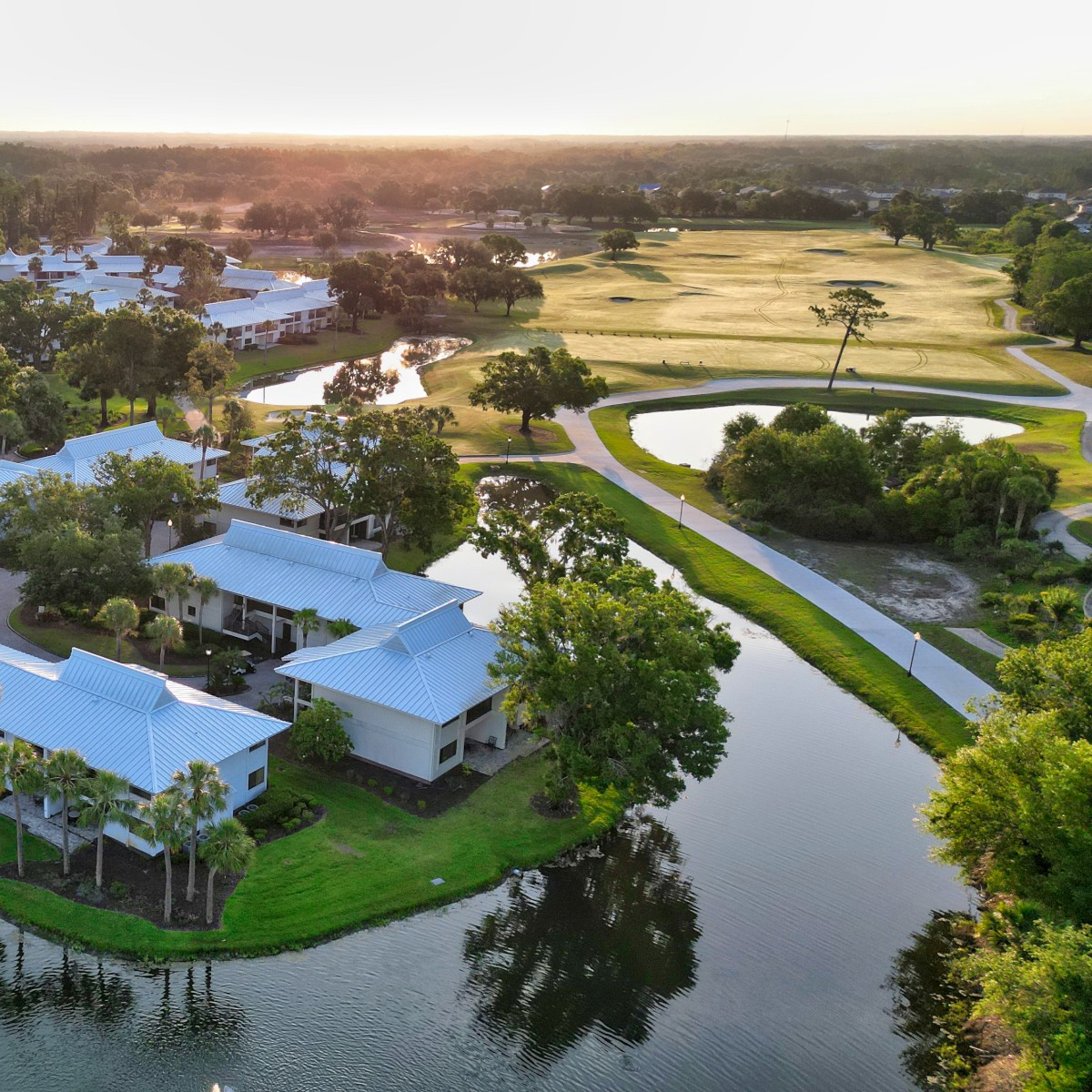 The image shows a scenic aerial view of a community with water channels, lush greenery, and buildings with white roofs at sunset.