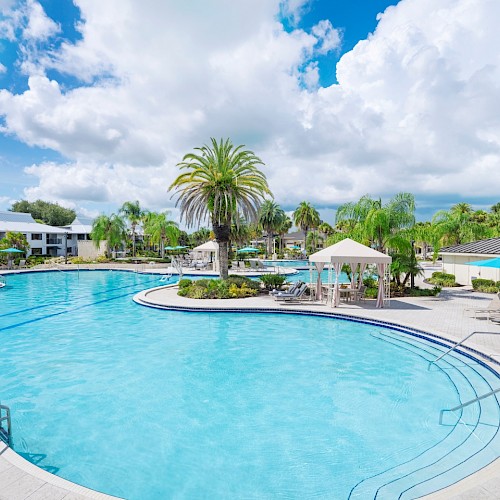 A sunny poolside setting with lounge chairs, palm trees, and cabanas. The sky is partly cloudy, creating a serene atmosphere.