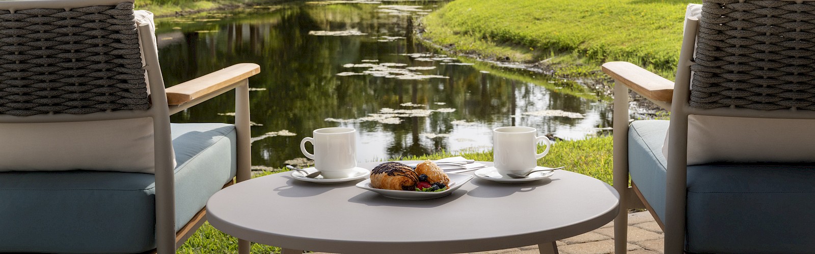 Two chairs face a scenic pond surrounded by greenery, with a table holding coffee cups and pastries in the foreground.
