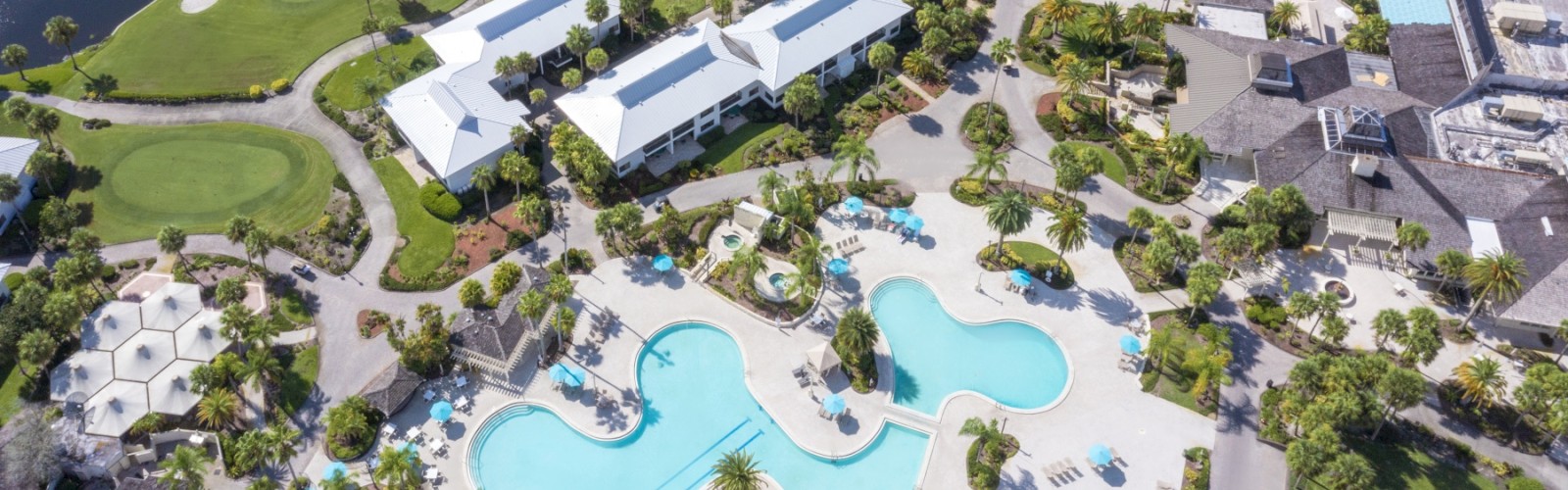 The image shows an aerial view of a resort featuring large swimming pools, white-roofed buildings, landscaped gardens, and water features.