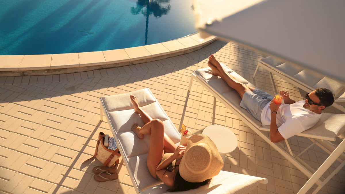 People lounging by a pool on sunbeds, a shaded umbrella overhead, relaxing vacation vibe by a bright tiled poolside.