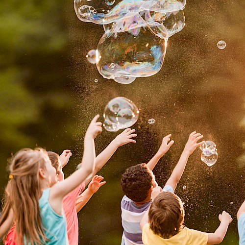 A group of children excitedly reaching for large, floating soap bubbles in an outdoor setting, captured against a blurred, green background.