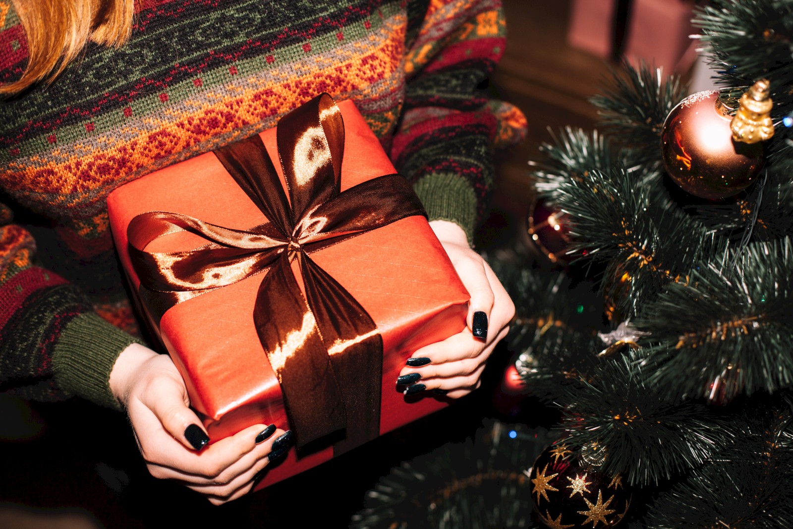 A person in a sweater holds a wrapped gift with a bow near a decorated Christmas tree with ornaments.