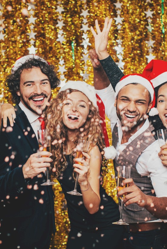 A group of five people wearing Santa hats and holding drinks, celebrating Christmas against a gold, festive backdrop with confetti falling.