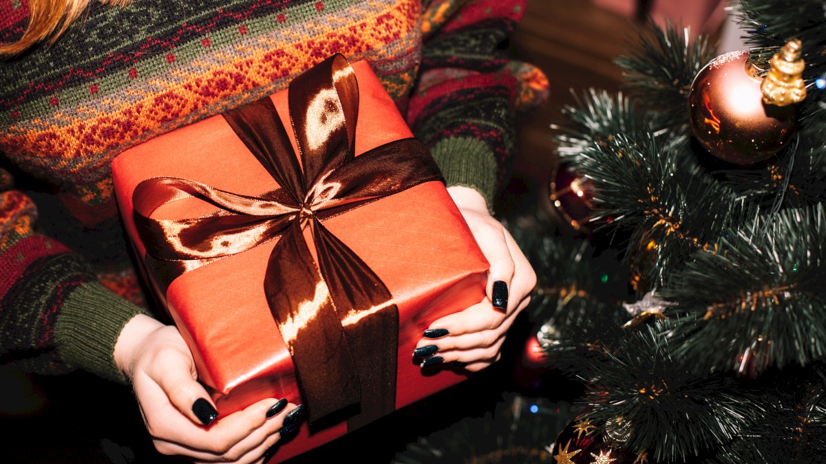 A person holds a wrapped gift with a brown ribbon near a decorated Christmas tree adorned with ornaments.