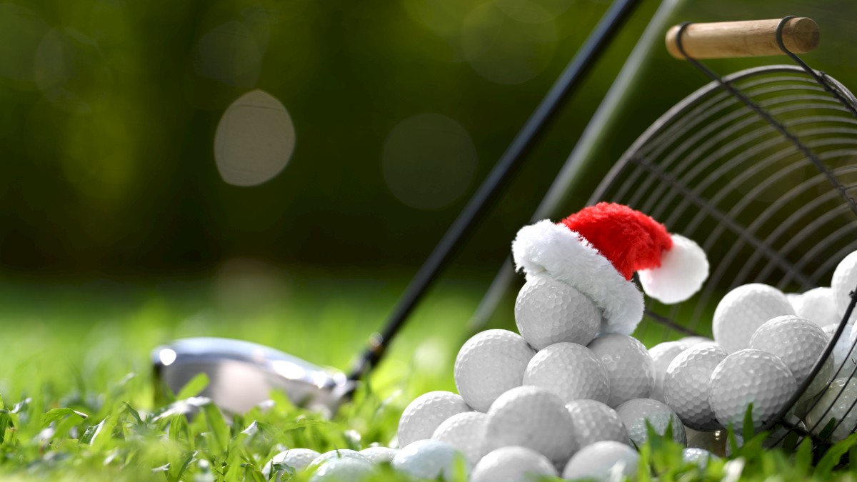 A tipped-over wire basket of golf balls, with one golf ball wearing a small Santa hat, alongside a golf club on a grassy field.