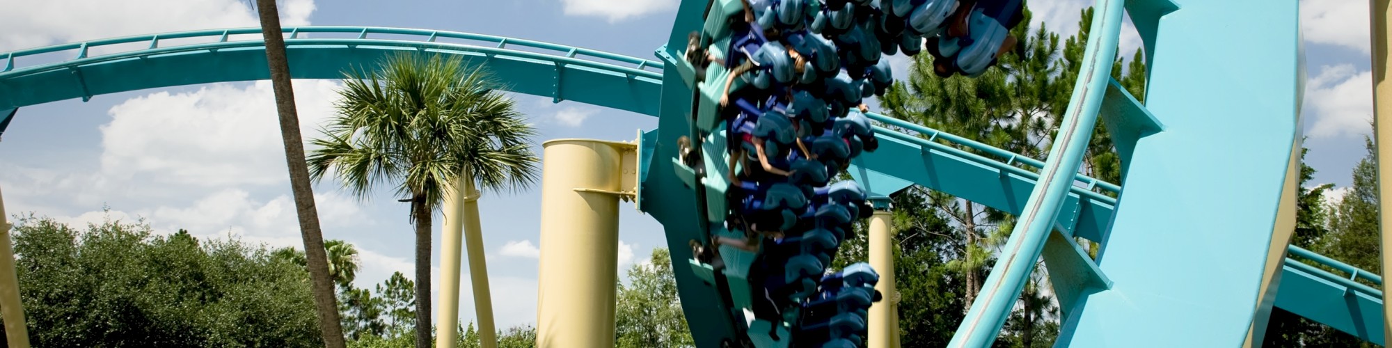 A turquoise roller coaster with riders is shown looping, surrounded by palm trees and greenery under a partly cloudy sky.