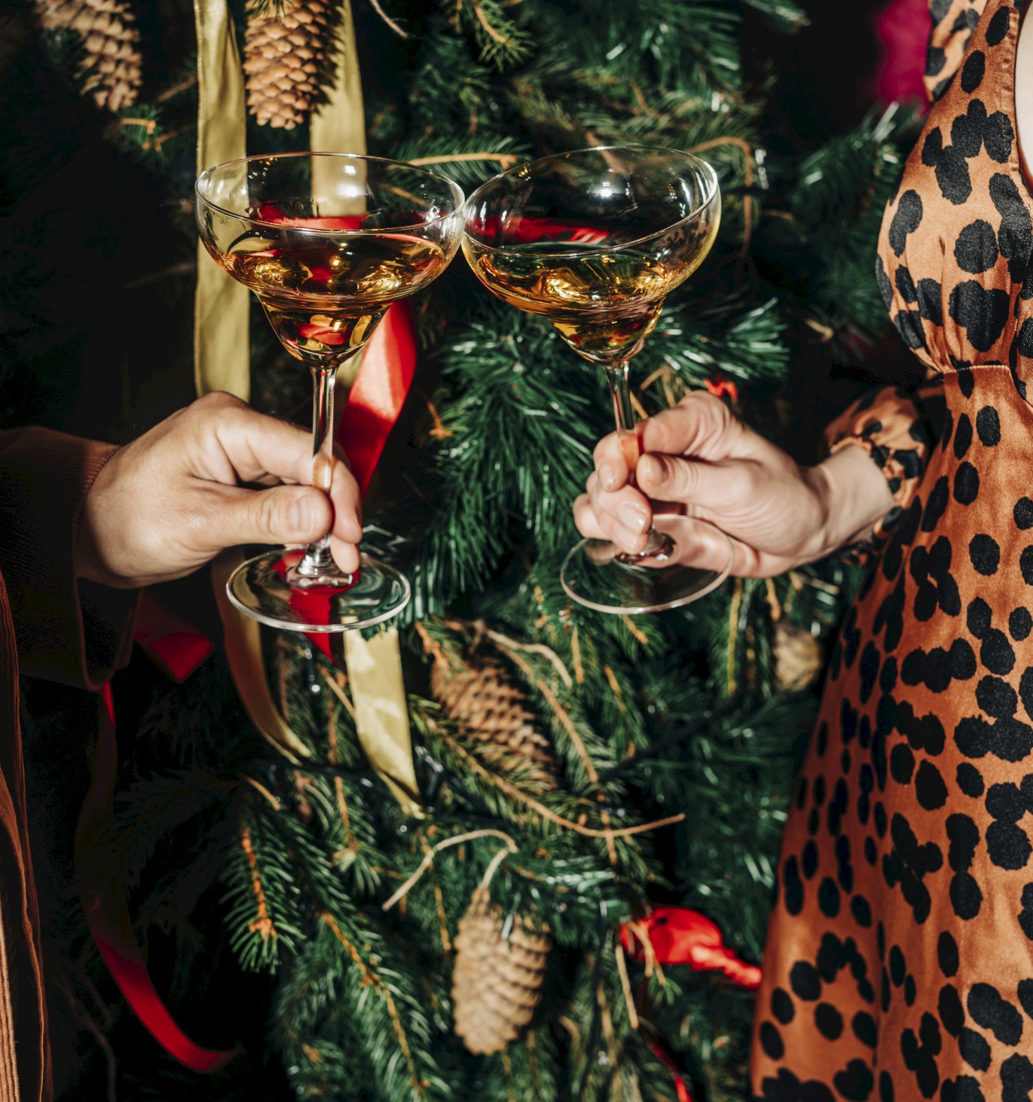 Two people clink champagne glasses in front of a decorated tree, wearing festive attire.
