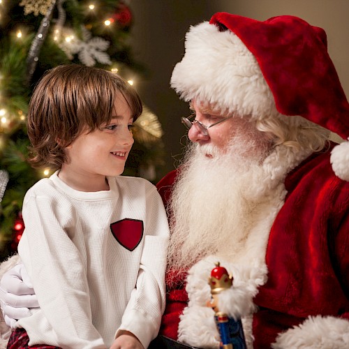 A child sits on Santa's lap beside a decorated Christmas tree, smiling and holding a toy in a festive setting.
