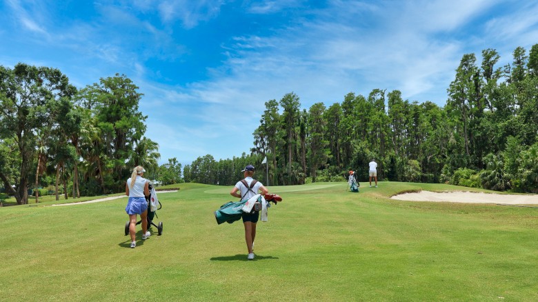 People are walking on a golf course carrying clubs, surrounded by green grass, trees, and a blue sky with clouds.