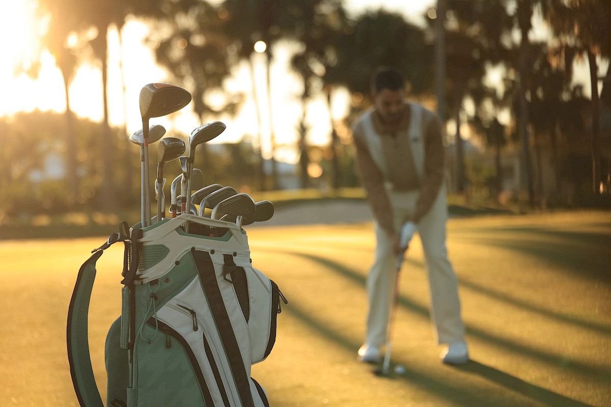 A golfer preparing to swing on a sunlit course, with a white golf bag and clubs in the foreground and palm trees in the background, warm evening light.