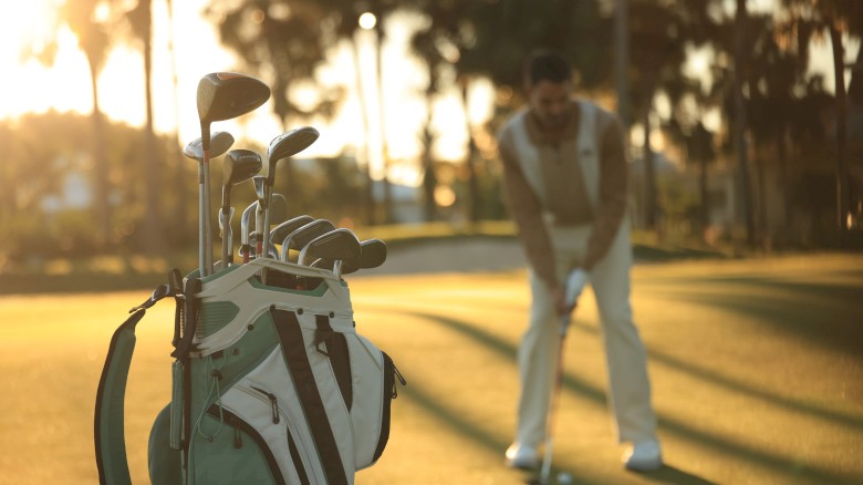 A golfer preparing to swing on a sunlit course, with a white golf bag and clubs in the foreground and palm trees in the background, warm evening light.