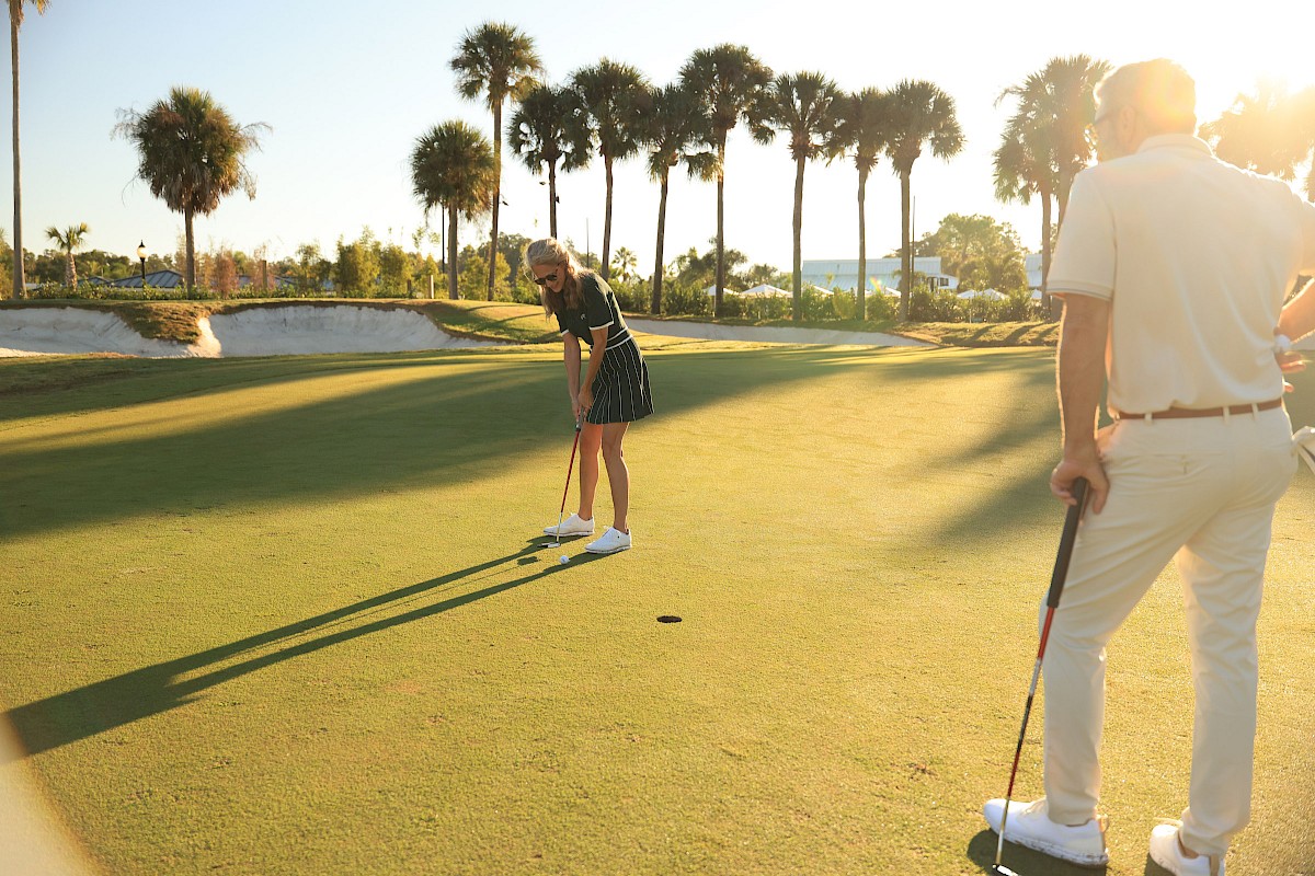 A golfer lines up a putt on a sunny course, while another player watches; palm trees frame the scene with long shadows.