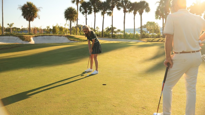 A golfer lines up a putt on a sunny course, while another player watches; palm trees frame the scene with long shadows.