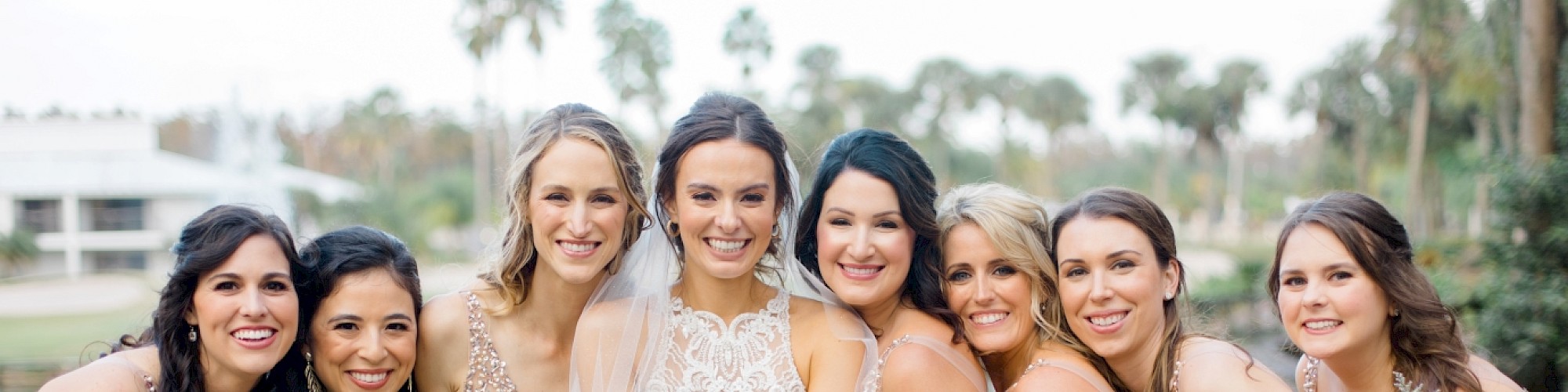 A bride and bridesmaids smiling, dressed in matching light peach dresses, holding colorful bouquets, outdoors with palm trees in the background.