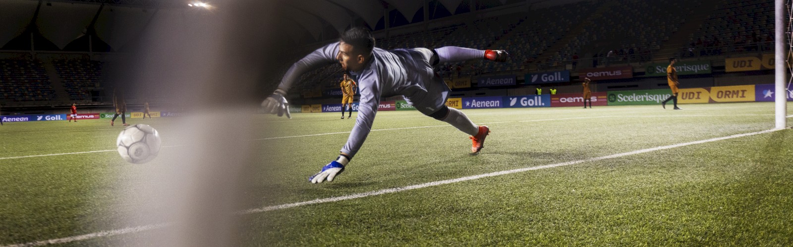 A goalkeeper in mid-air, attempting to save a soccer ball, under stadium lights, with the net partially visible in the foreground.