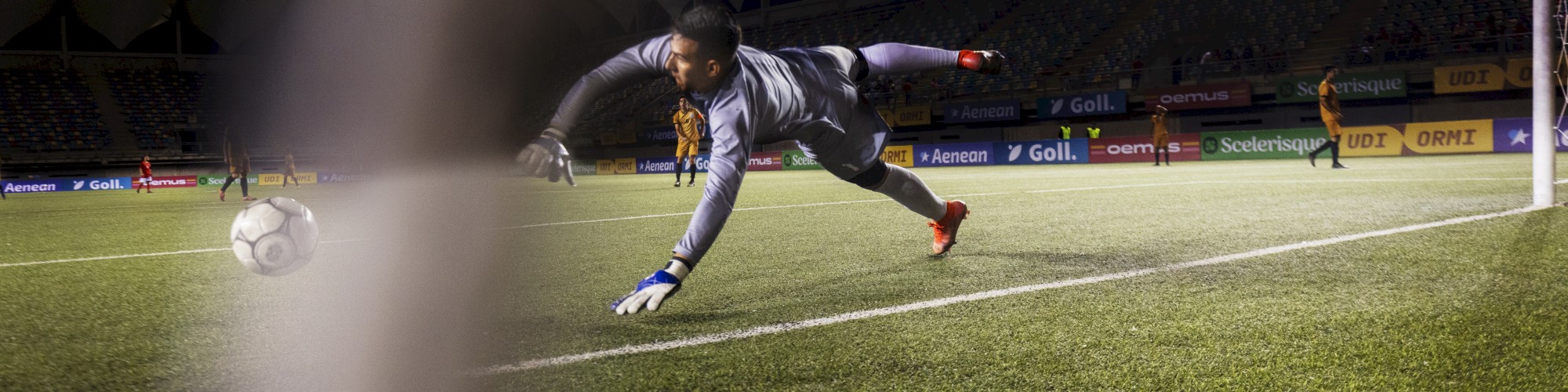 A goalkeeper in mid-air, attempting to save a soccer ball, under stadium lights, with the net partially visible in the foreground.