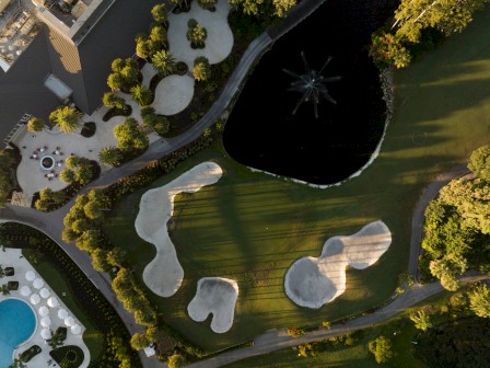 An aerial view of a landscaped park with winding paths, green lawns, trees, water features, and irregularly shaped white sand/stone patches.
