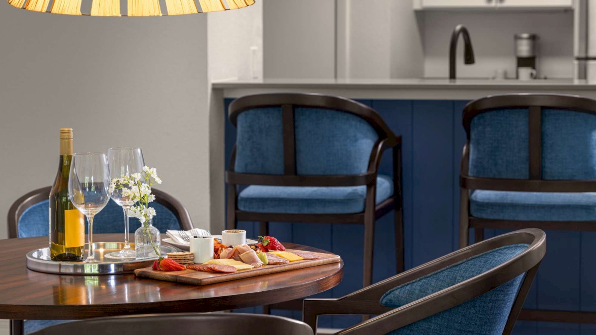 A cozy dining setup with wine, glasses, and a cheese platter on a wooden table; background shows a kitchen and blue bar stools.