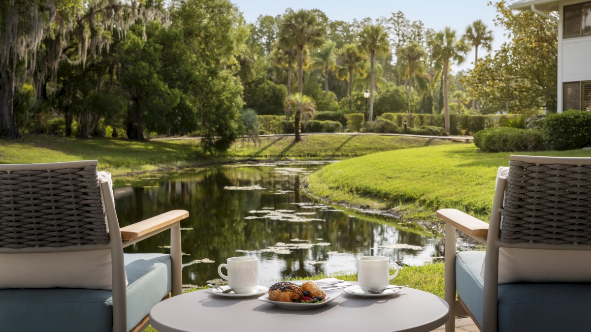 Two chairs on a patio overlook a tranquil pond, with two cups and a plate of pastries on a round table between them.