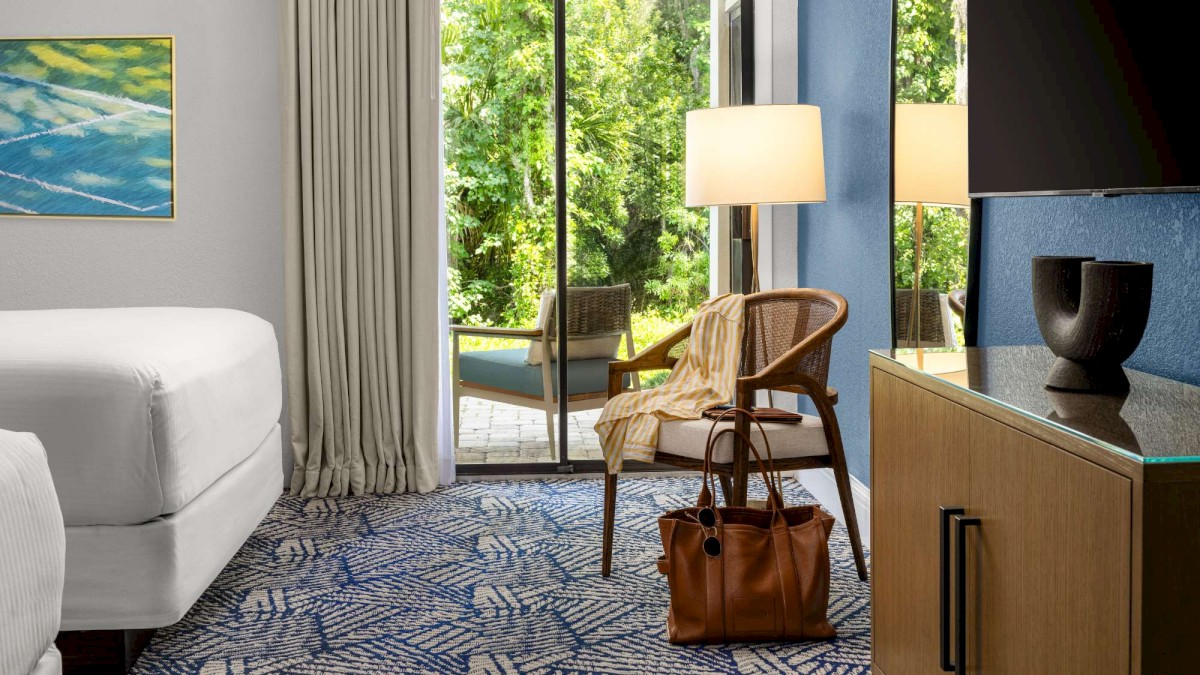 A modern hotel room with a bed, chair, lamp, and a view of greenery outside through a glass door. A decorative vase sits on a dresser.