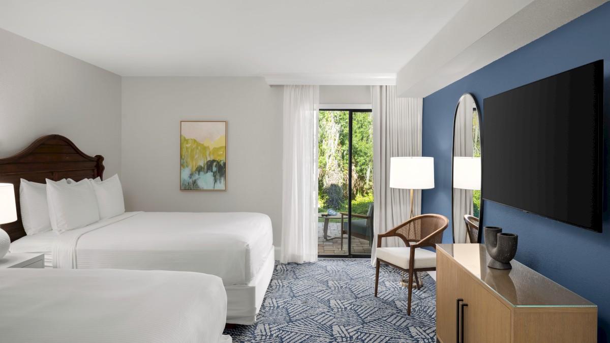A hotel room with two beds, a TV, a blue accent wall, a chair, a lamp, art on the wall, and a window overlooking greenery.