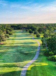 A lush golf course landscape with a winding path, trees, and a small lake under a clear blue sky stretches into the distance.