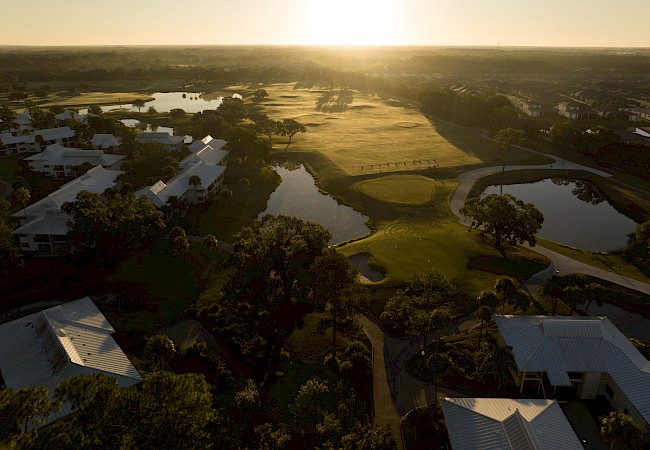 An aerial view of a golf course at sunrise, with lush greenery, water features, and surrounding buildings.