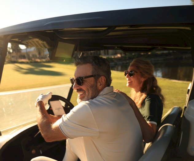 A couple in a golf cart enjoying a sunny day outdoors, wearing sunglasses and smiling as they ride through the green landscape.