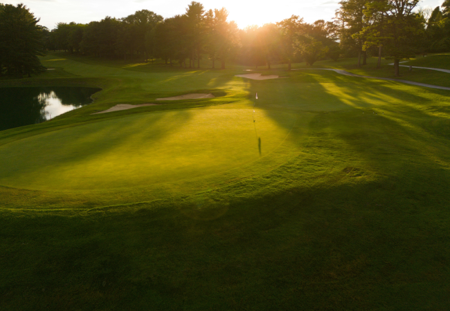 A serene golf course at sunset, with sunlight casting long shadows on the green grass and a water hazard nearby.