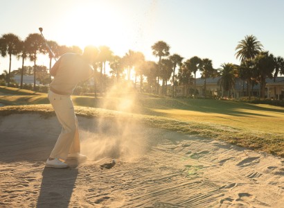 A person is hitting a golf ball out of a sand trap on a sunny course with palm trees in the background.