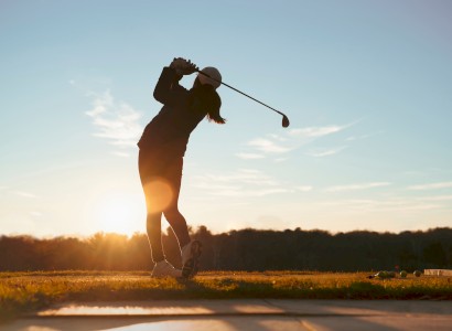 A person is playing golf at sunset, captured mid-swing, with a scenic landscape in the background.