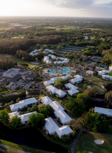 An aerial view of a resort complex with buildings, pools, and greenery surrounded by a vast landscape, under a partially cloudy sky.