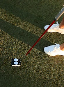 A person is playing golf, wearing white sneakers and a skirt, holding a red putter, about to putt a ball on the green.