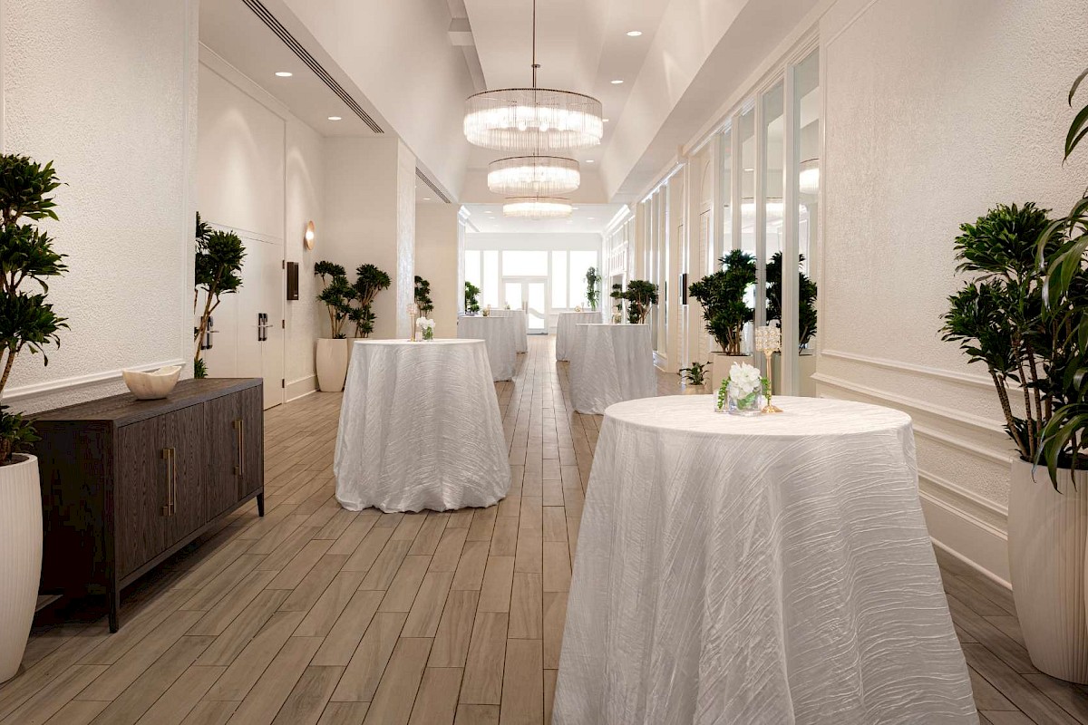 An elegant hallway with tall plants, round tables with white tablecloths, and modern chandeliers. Natural light filters through the windows.