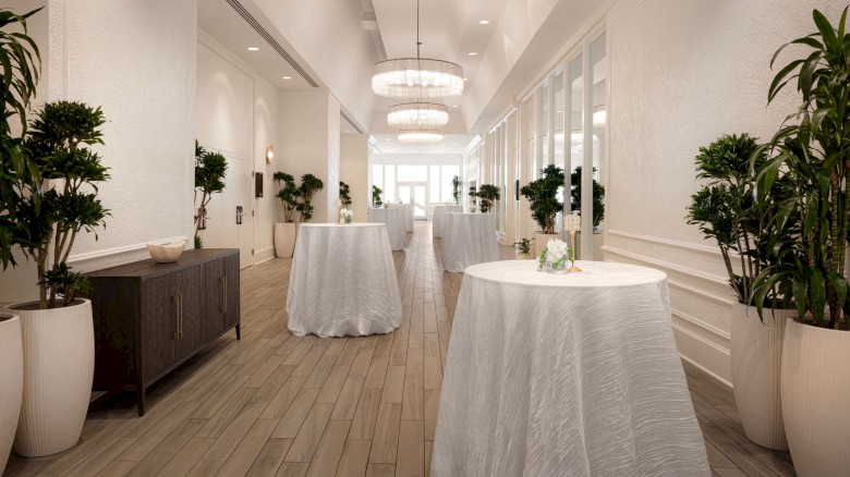 An elegant hallway with tall plants, round tables with white tablecloths, and modern chandeliers. Natural light filters through the windows.