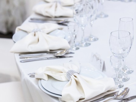 A neatly arranged dining table with white napkins, glassware, and silver cutlery on a white tablecloth, set for a formal event.