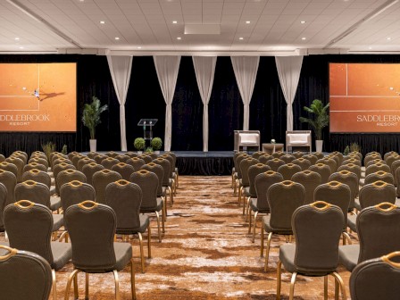 An empty conference room with rows of chairs facing a stage, two screens showing 