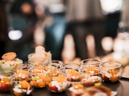 The image shows a table with small dessert cups in the foreground, with people socializing in a blurred background.