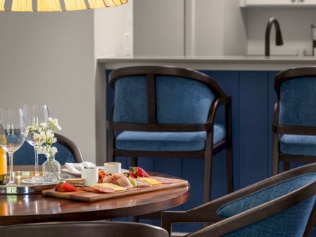 A cozy dining setup with wine, glasses, and a cheese platter on a table, two blue chairs, and a kitchen island in the background.