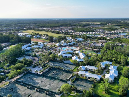 The image shows an aerial view of a residential area with tennis courts, houses, trees, and roads surrounded by greenery and open land.